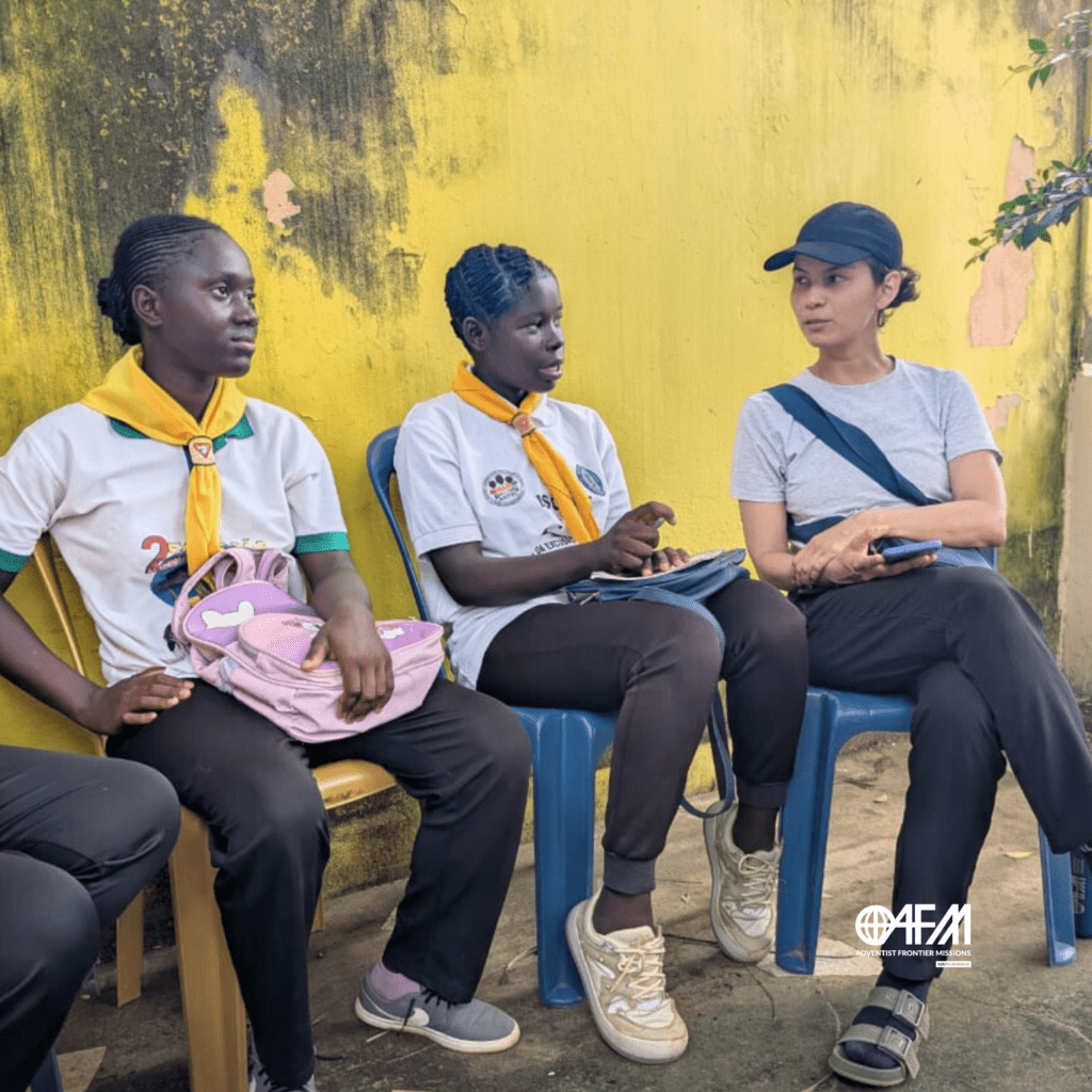 Kuma ku bu sta - missionária sentada ao lado de jovens guineenses conversando.